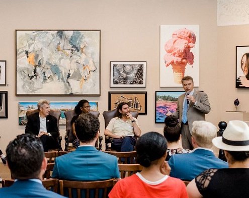 man speaking to crowd beside panel with art in the background